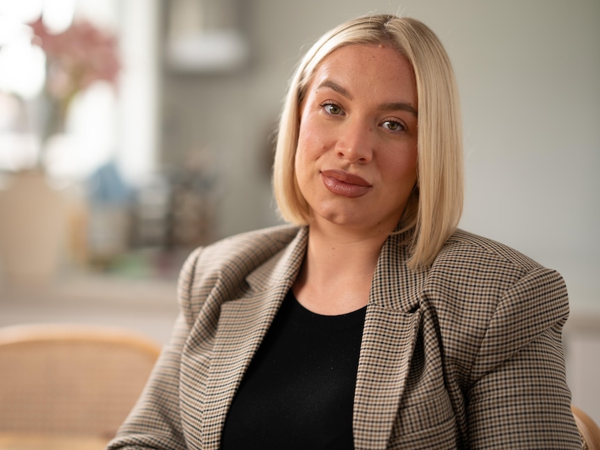 a young blonde woman wearing a tweed blazer over a black top looking serious