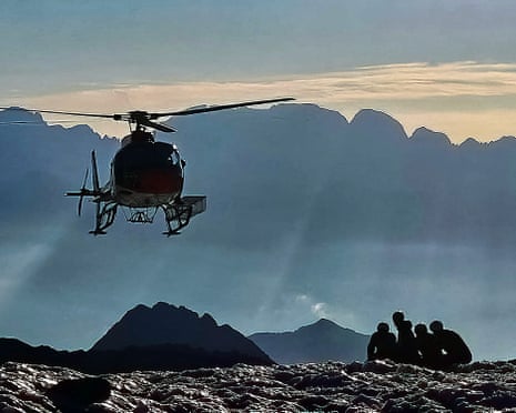 A small group of soldiers crouched on the ground as a helicopter flies nearby against a backdrop of mountain peaks