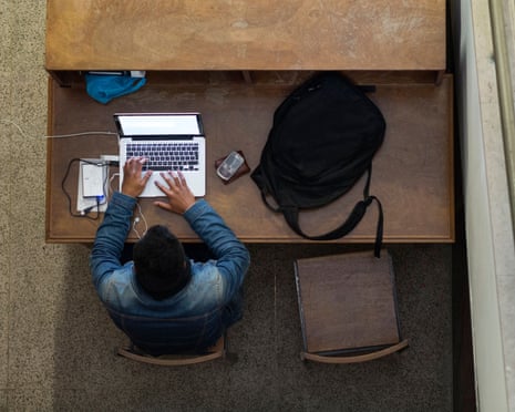 Overhead view of a man typing on a laptop at a wooden desk with a backpack