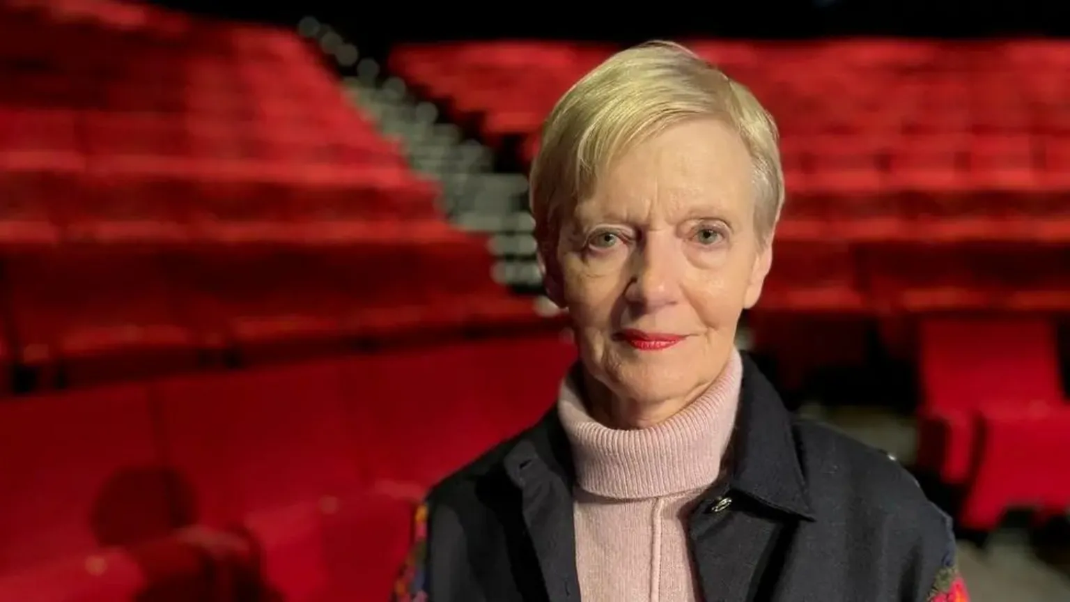 BBC A woman with a serious expression looking into the camera in a theatre with red seats stretching out in the auditorium behind her
