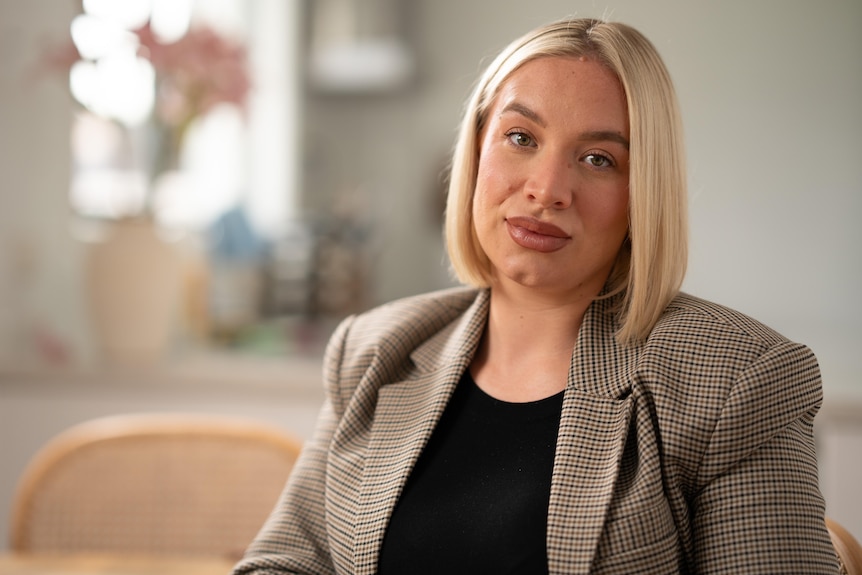 a young blonde woman wearing a tweed blazer over a black top looking serious